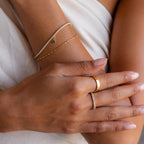 Close-up of hands wearing gold and diamond bracelets—featuring the Heart Birthstone Tennis Bracelet—and personalized charm rings, resting on light fabric.
