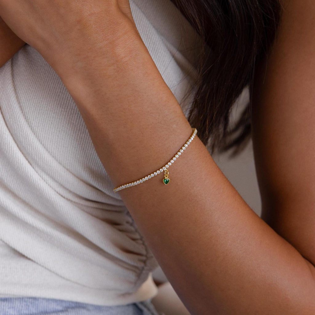 A woman wears the Heart Birthstone Tennis Bracelet, featuring a gold band with a small green heart charm, paired with a sleeveless white top.