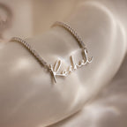 A Diamond Tennis Name Necklace with a personalized pendant is displayed on a smooth, cream-colored surface.