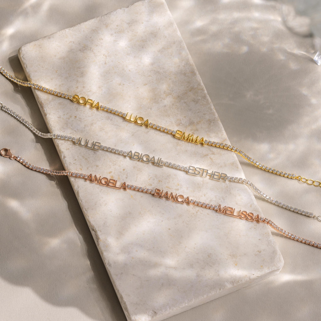 The Multiple Name Tennis Bracelet, available in gold, silver, and rose gold with custom name charms, is elegantly displayed on a cream stone slab.