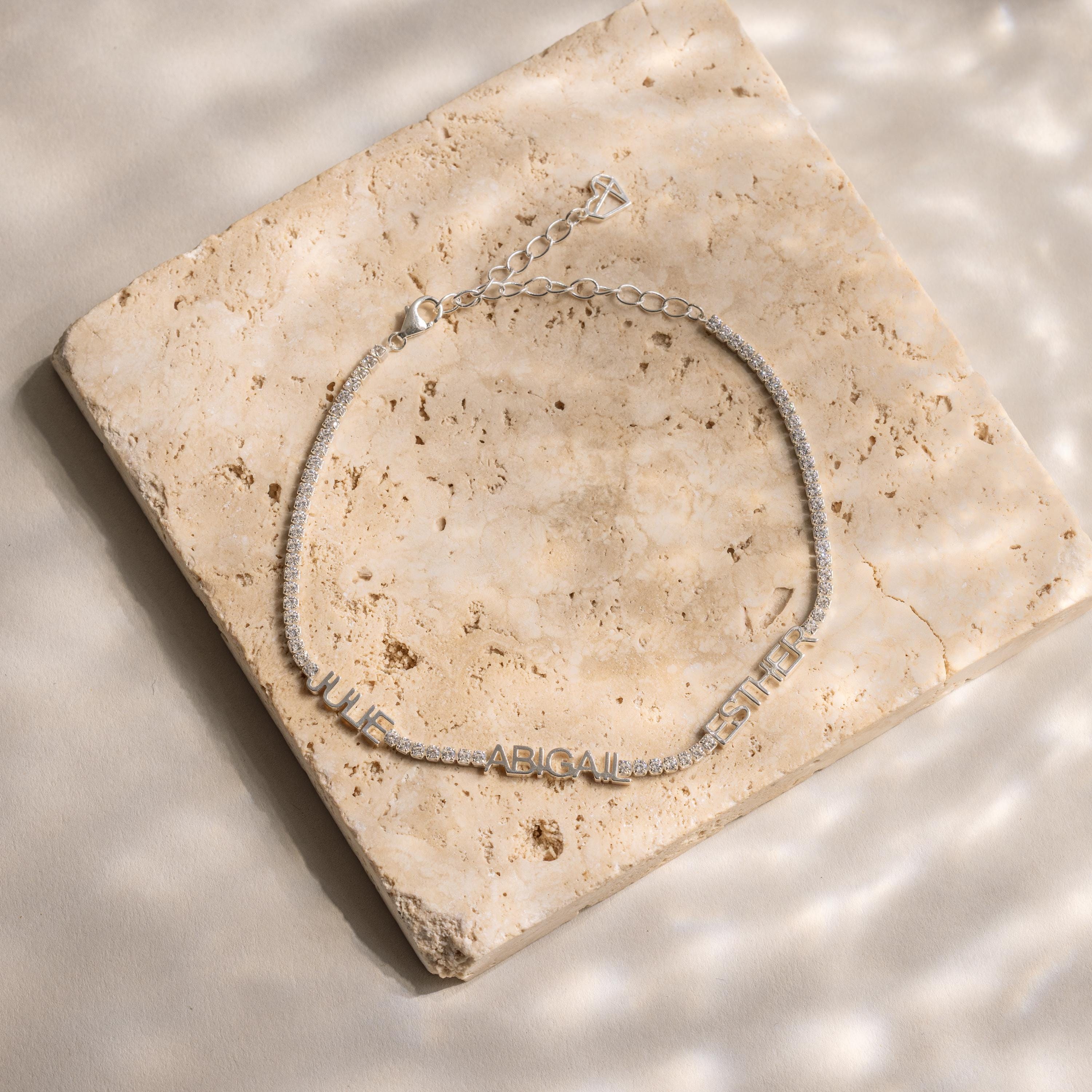 A silver Multiple Name Tennis Bracelet featuring letter charms spelling JULIE ABIGAIL ESTHER rests on a square marble tile.