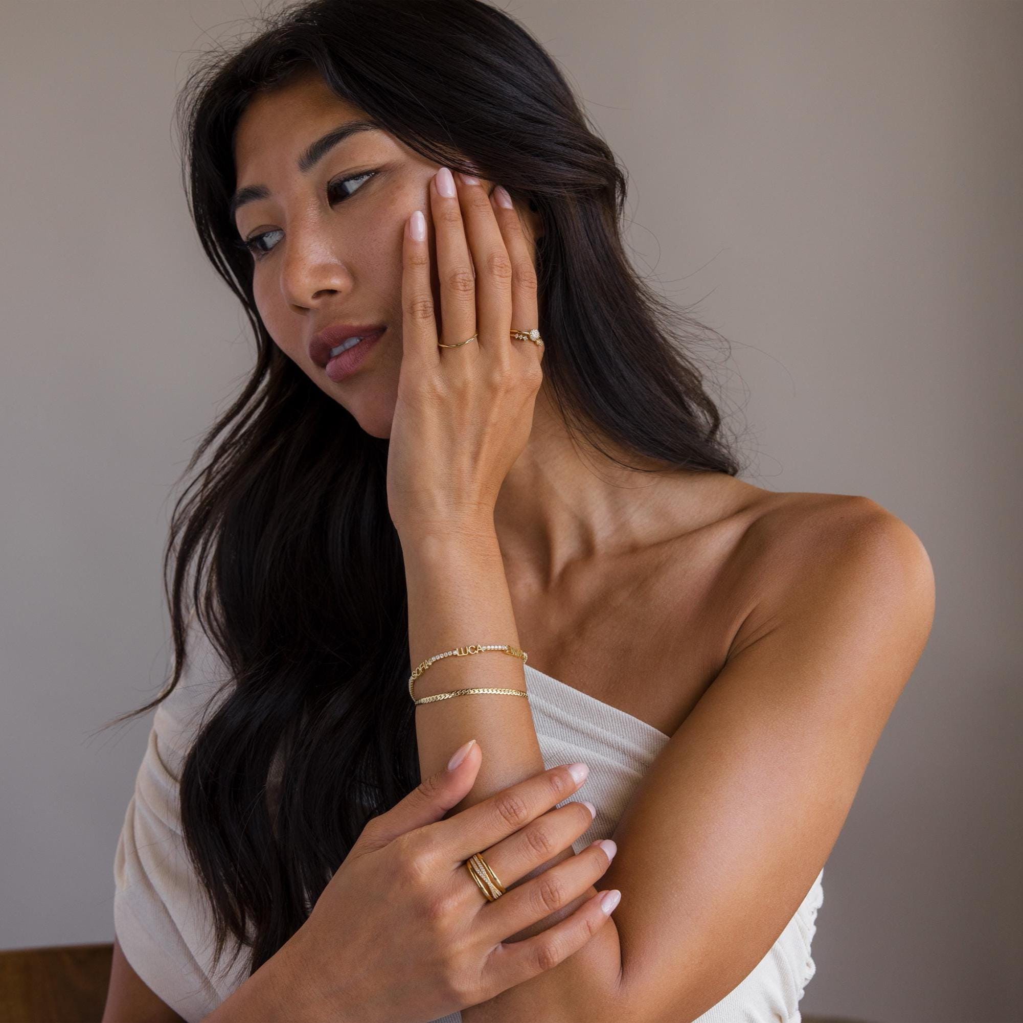 A woman with long dark hair, in an off-shoulder top, touches her face and chest while wearing gold jewelry, including the Multiple Name Tennis Bracelet.