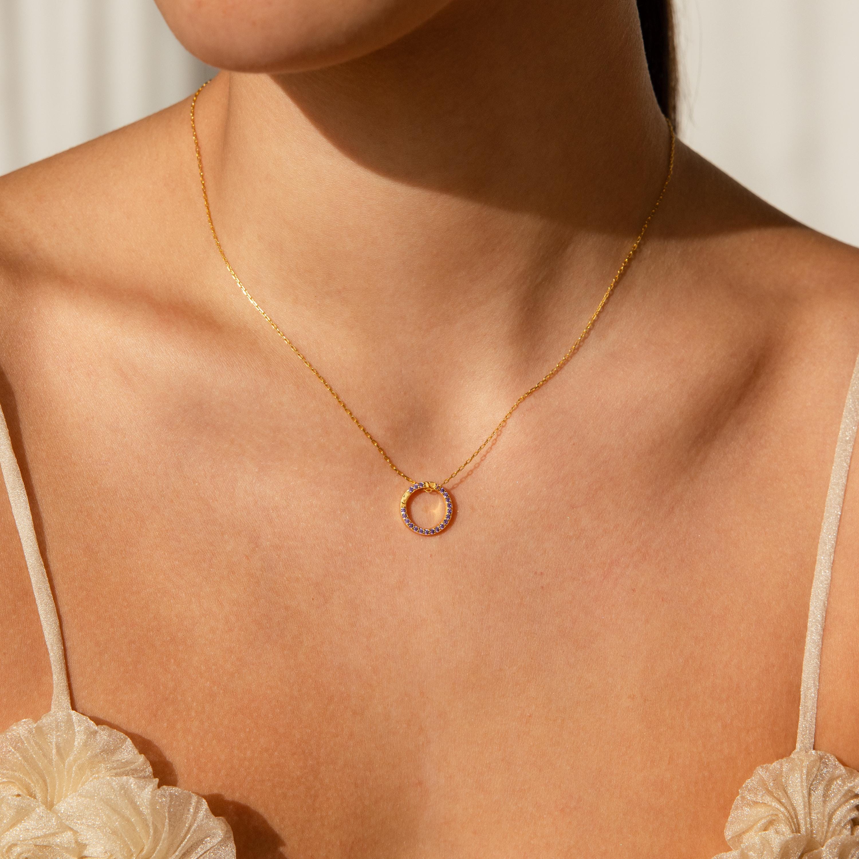 A woman wearing a gold necklace with a small circular pendant, dressed in a cream-colored top—perfect for pairing with customizable jewelry like the Pave Amethyst Charm Clip.