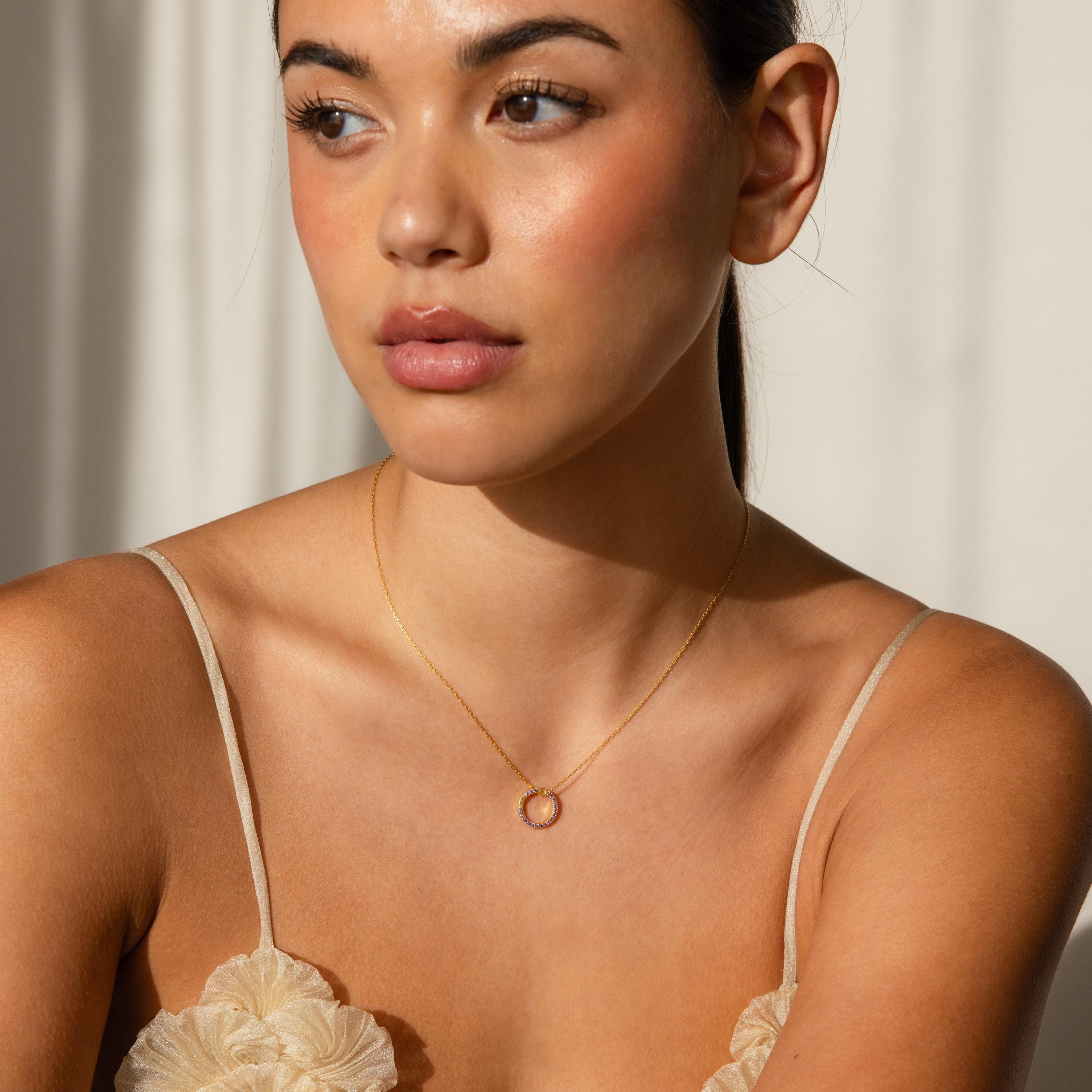 Woman wearing the Pave Amethyst Charm Clip, a gold necklace with a circular pendant, looking to the side in soft natural light—an elegant example of customizable jewelry.