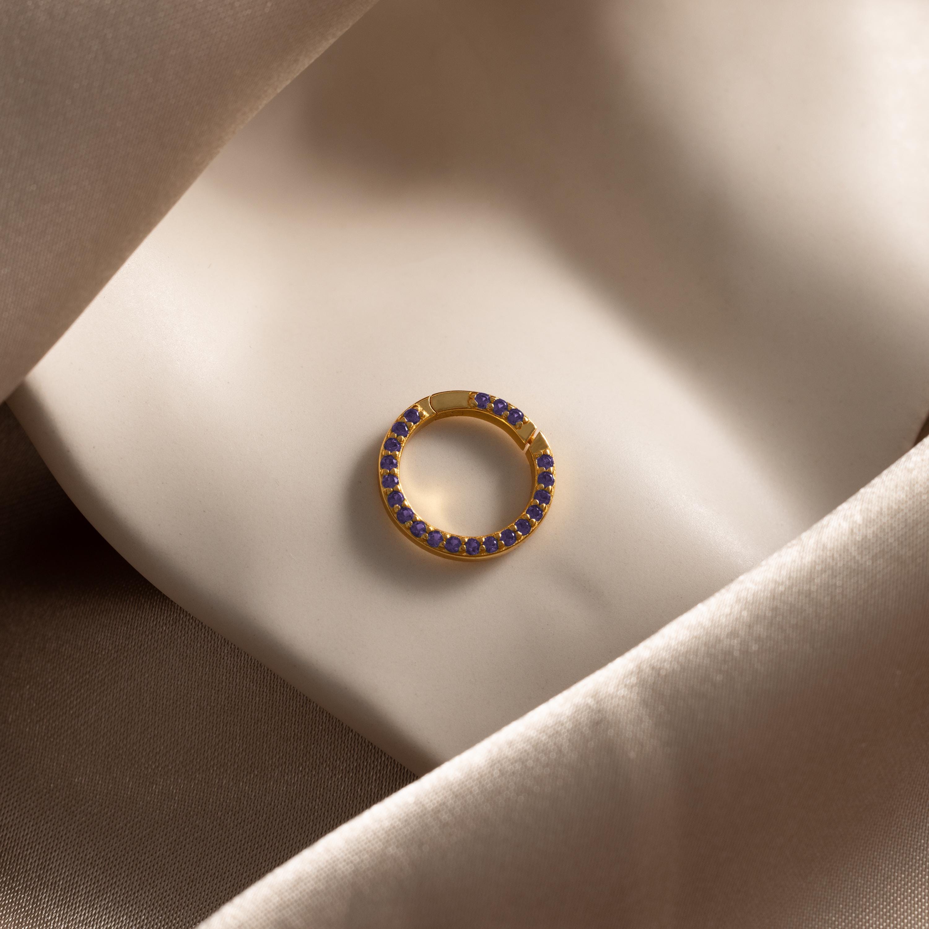 The Pave Amethyst Charm Clip, a gold circular earring adorned with purple gemstones, is displayed on smooth beige satin fabric.