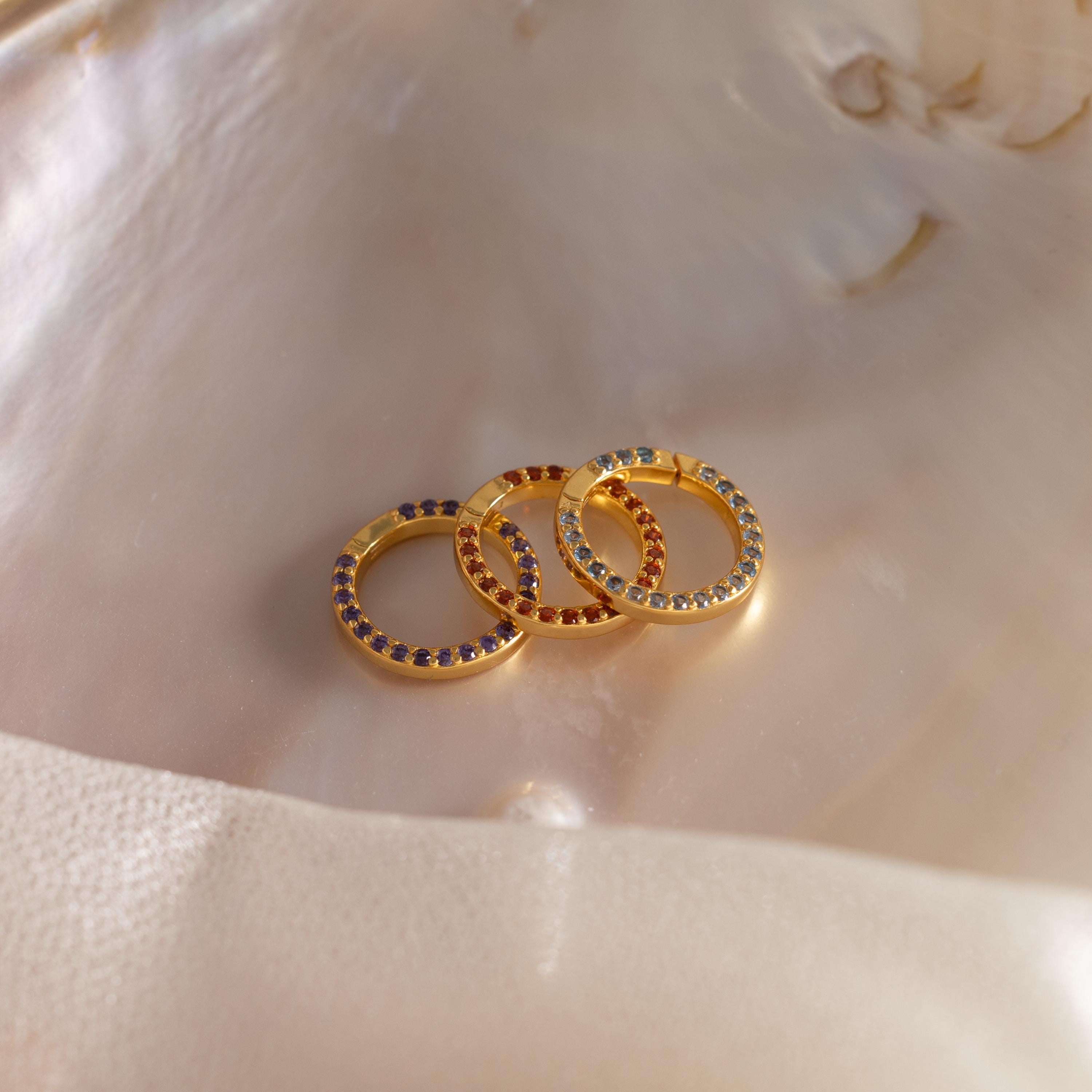 Three gold rings, including a Pave Aquamarine Charm Clip with shimmering aquamarine stones, are displayed on a shiny, iridescent shell surface.