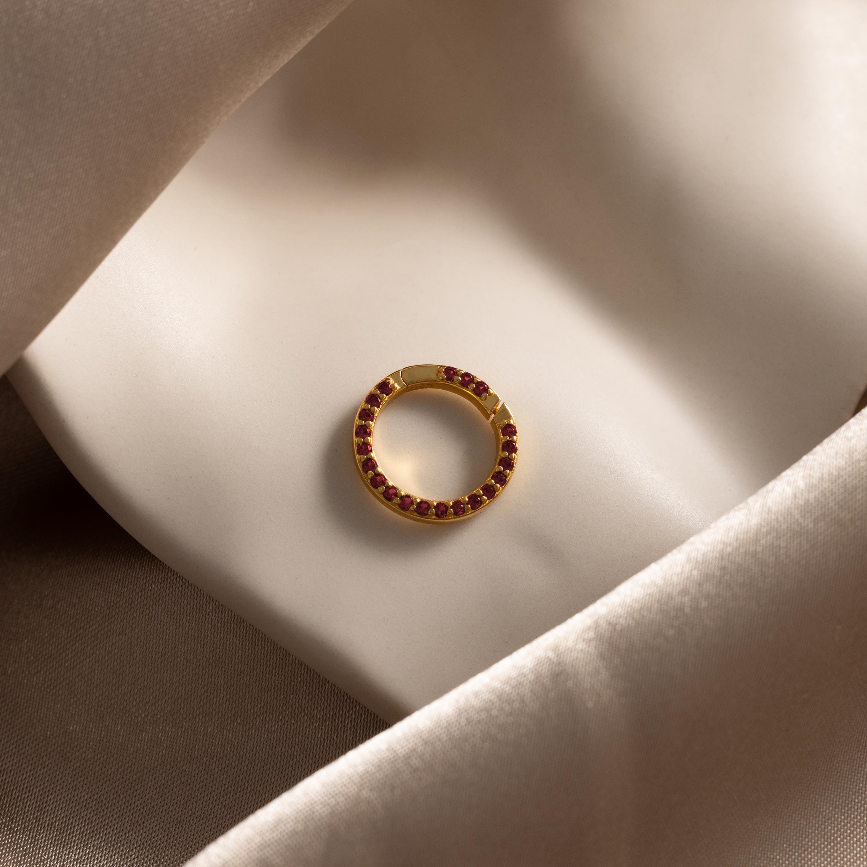 The Pave Garnet Charm Clip, featuring gold and red gemstones, rests on folded champagne satin—ideal for personalizing your jewelry.