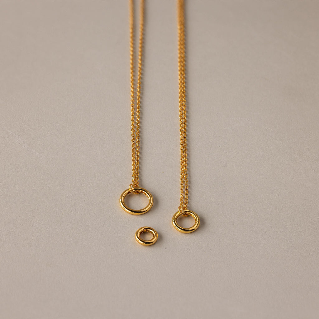 The Round Carabiner Charm Set in 18K Gold features two elegant gold necklaces with circular pendants and a loose ring, perfect for layering or pairing with your favorite charm necklace on a neutral background.