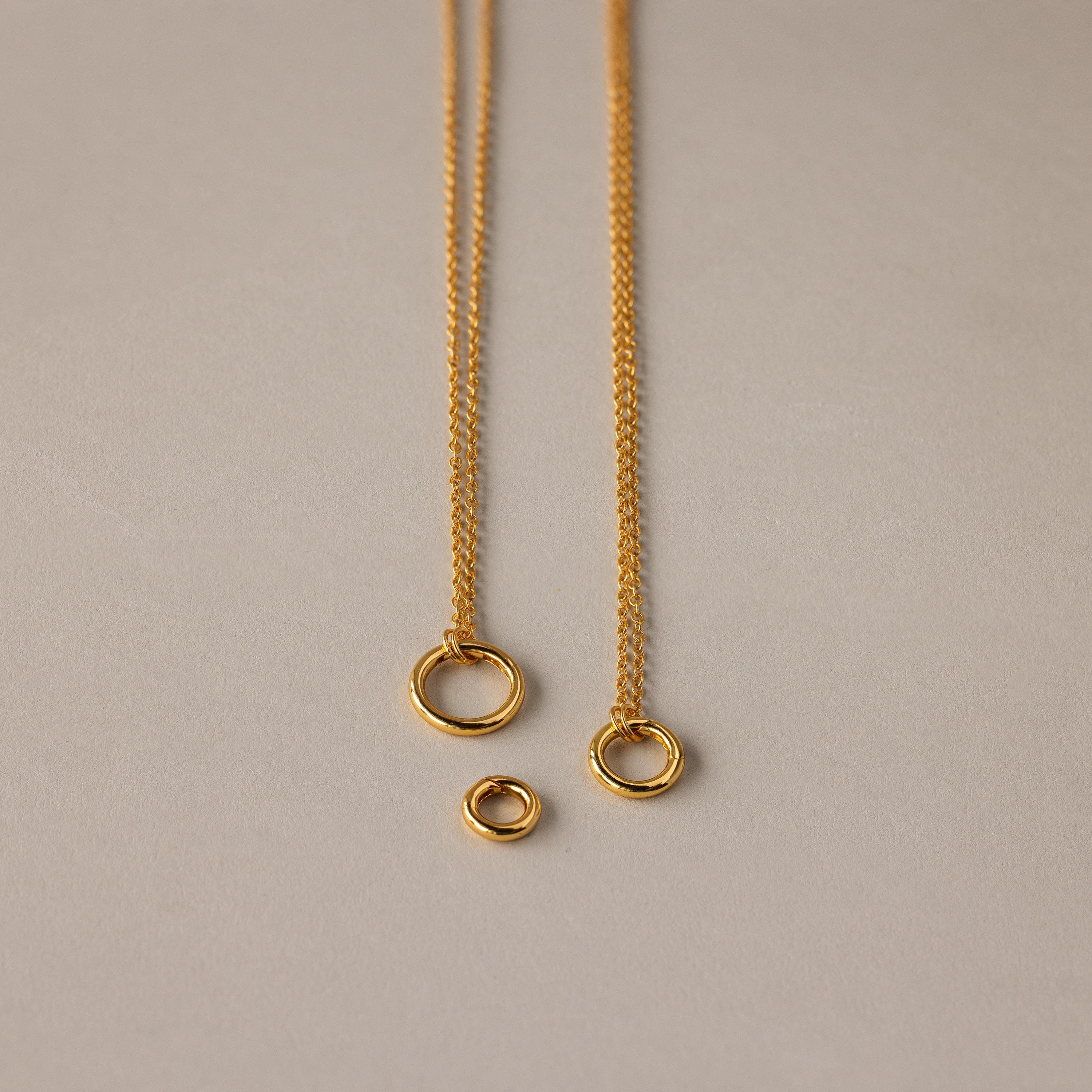 The Round Carabiner Charm Set in 18K Gold features two elegant gold necklaces with circular pendants and a loose ring, perfect for layering or pairing with your favorite charm necklace on a neutral background.