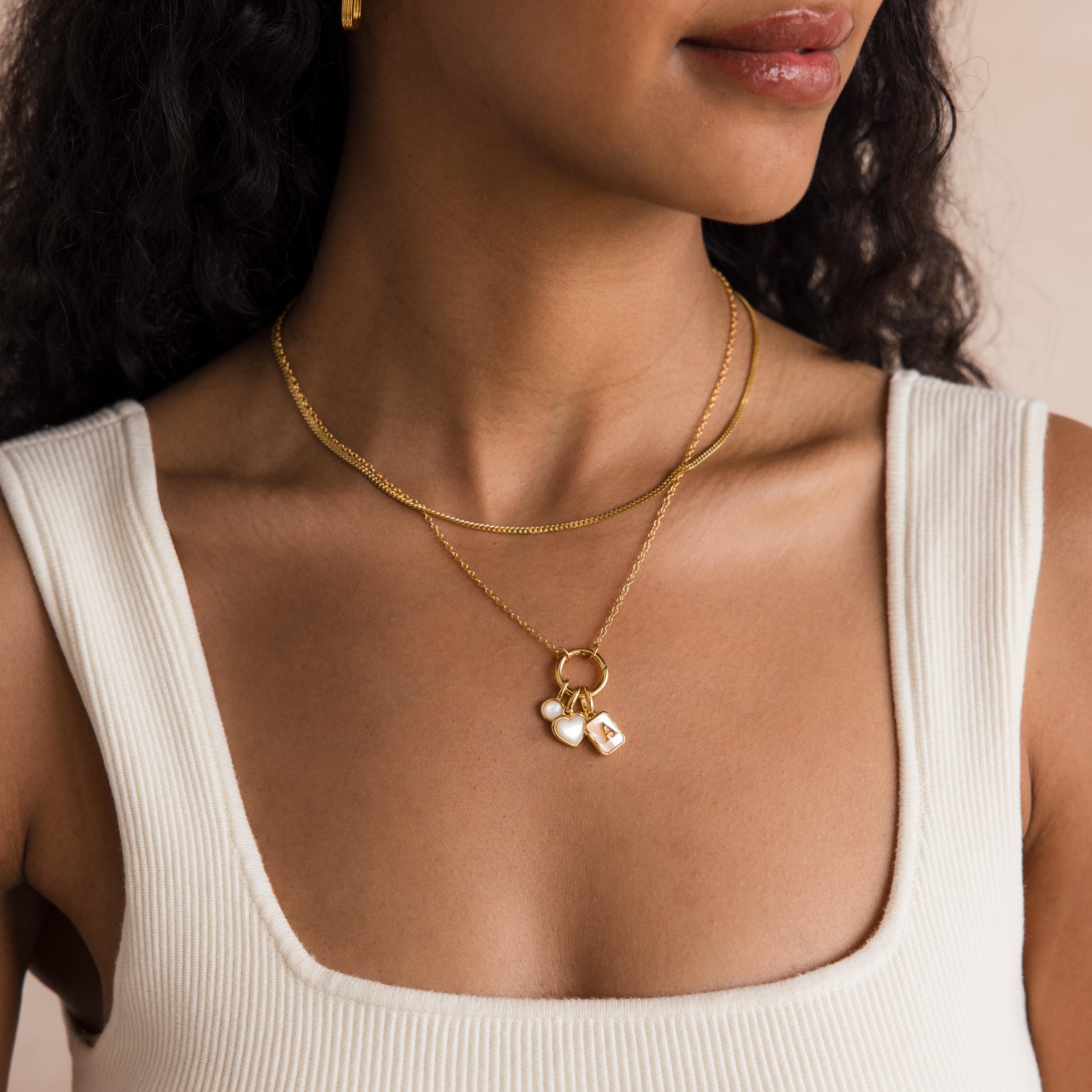 A woman wears a white sleeveless top and layered gold necklaces, featuring letter and heart pendants along with the Round Carabiner Charm Set in 18K Gold.