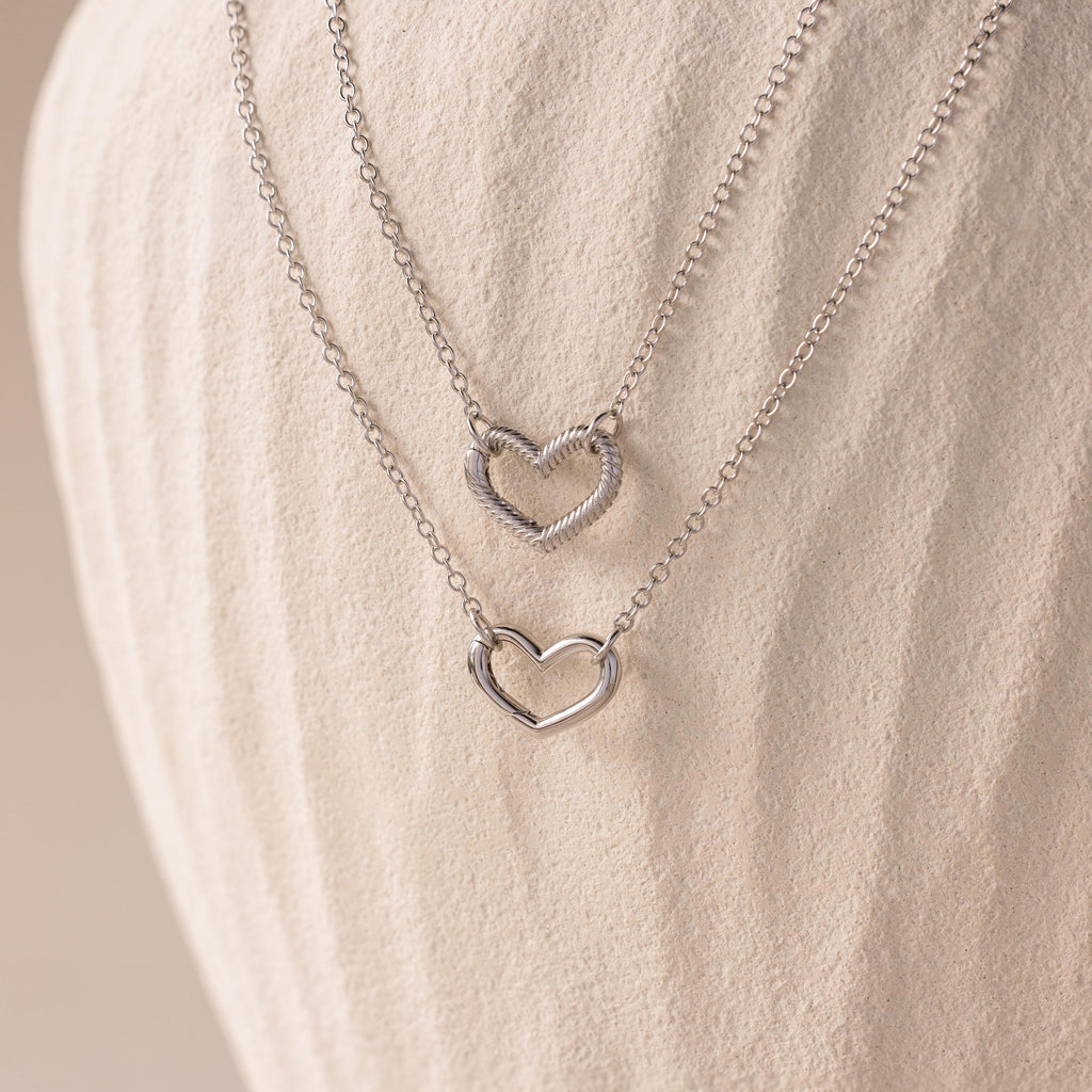 The Heart Carabiner Set in Sterling Silver features two heart-shaped pendant necklaces, ideal for layering with other jewelry, displayed on a textured, off-white background.