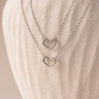 The Heart Carabiner Set in Sterling Silver features two heart-shaped pendant necklaces, ideal for layering with other jewelry, displayed on a textured, off-white background.