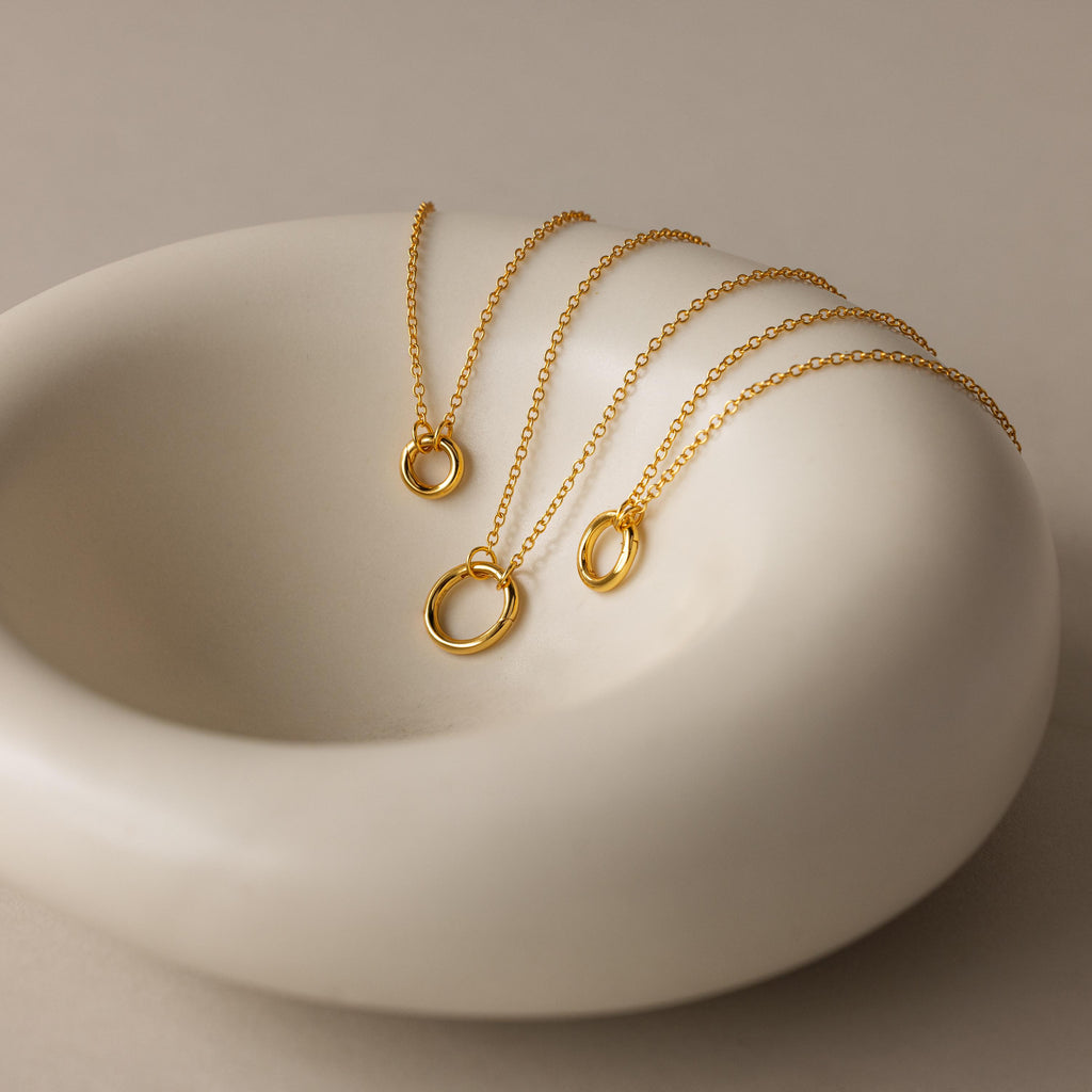 The Round Carabiner Charm Set in 18K Gold, featuring circular pendants, is displayed on a smooth, white ceramic dish.