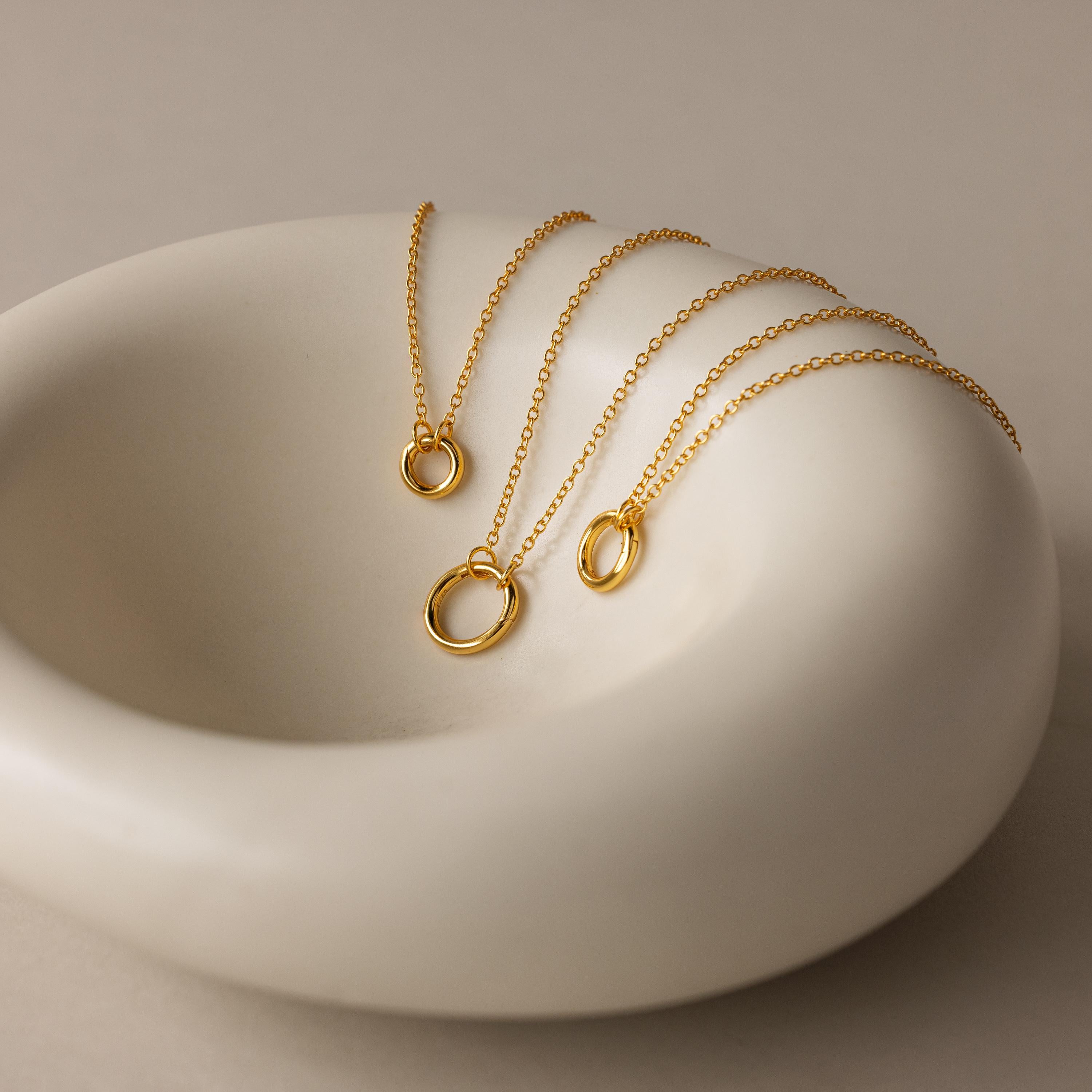 The Round Carabiner Charm Set in 18K Gold, featuring circular pendants, is displayed on a smooth, white ceramic dish.