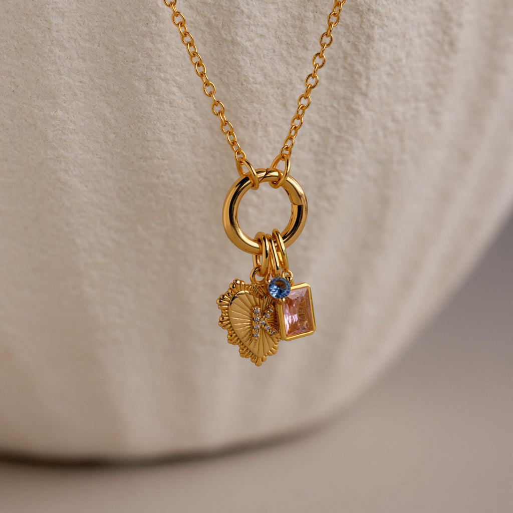 The Cable Circle Charm Necklace features three statement charms—a heart, a blue gem, and a pink square gem—on a gold chain, displayed against a neutral background. This elegant necklace enhances any outfit.