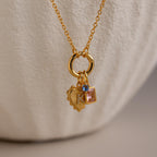 The Cable Circle Charm Necklace features three statement charms—a heart, a blue gem, and a pink square gem—on a gold chain, displayed against a neutral background. This elegant necklace enhances any outfit.