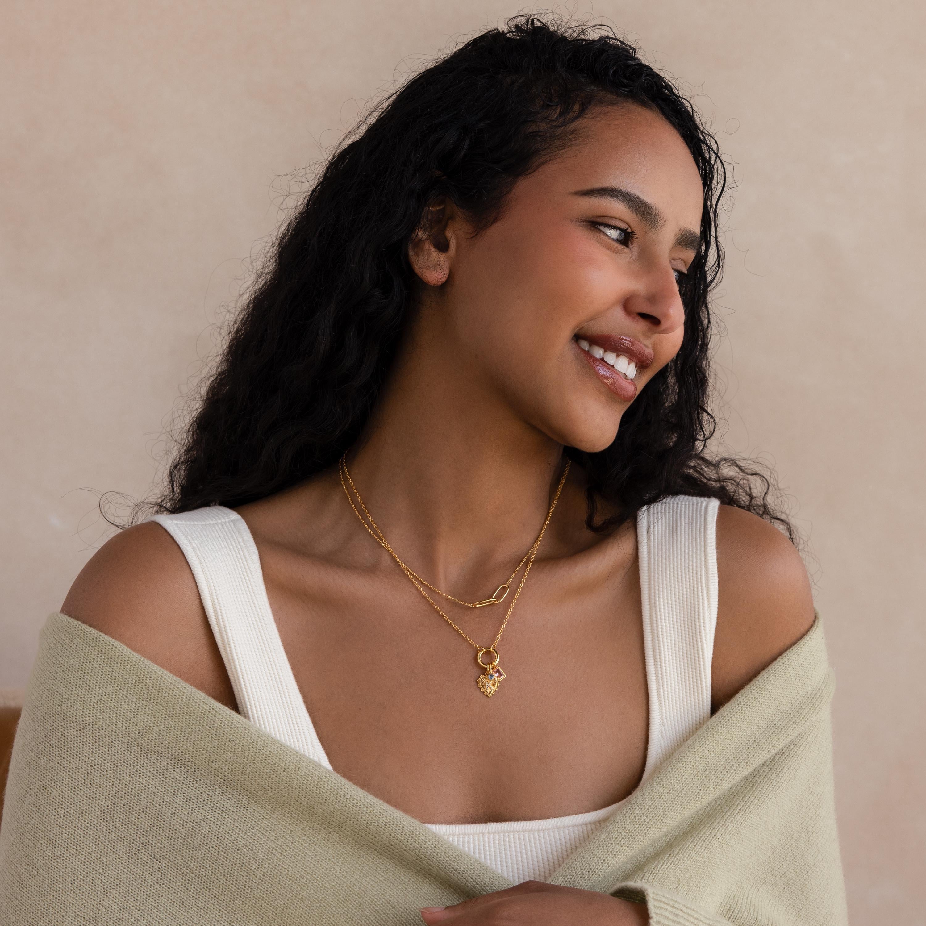 A smiling woman with long curly hair layers the Cable Circle Charm Necklace with other gold jewelry, pairing them with a cream shawl over a white top.