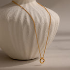 The Cable Circle Charm Necklace features a simple gold chain with a circular pendant, displayed on a white textured bust—an elegant and subtle statement piece.