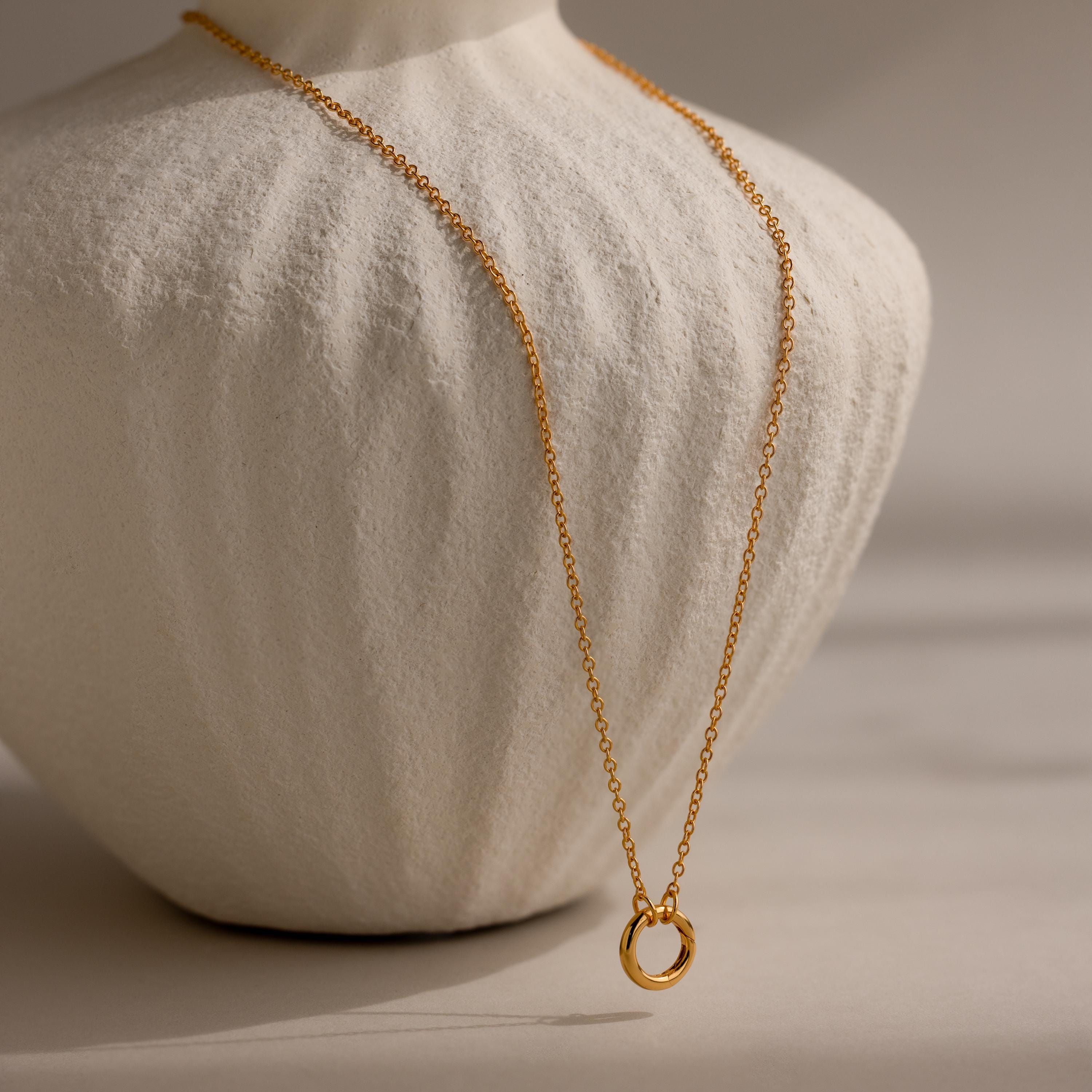 The Cable Circle Charm Necklace features a simple gold chain with a circular pendant, displayed on a white textured bust—an elegant and subtle statement piece.