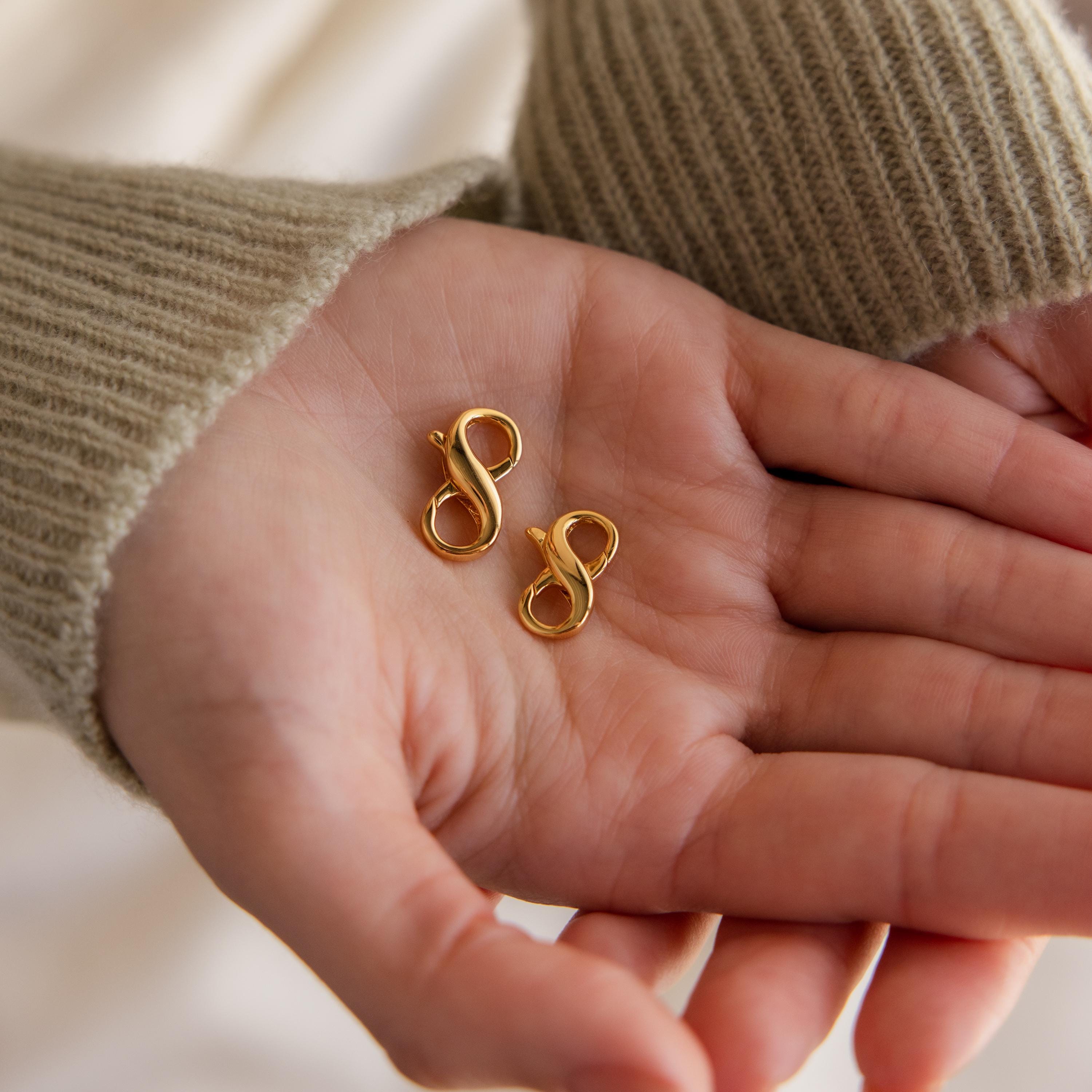 A hand holds two gold infinity earrings, ideal to pair with the Multipurpose Lobster Clasp Extender Set in 18K Gold for added versatility with bracelets or necklaces.