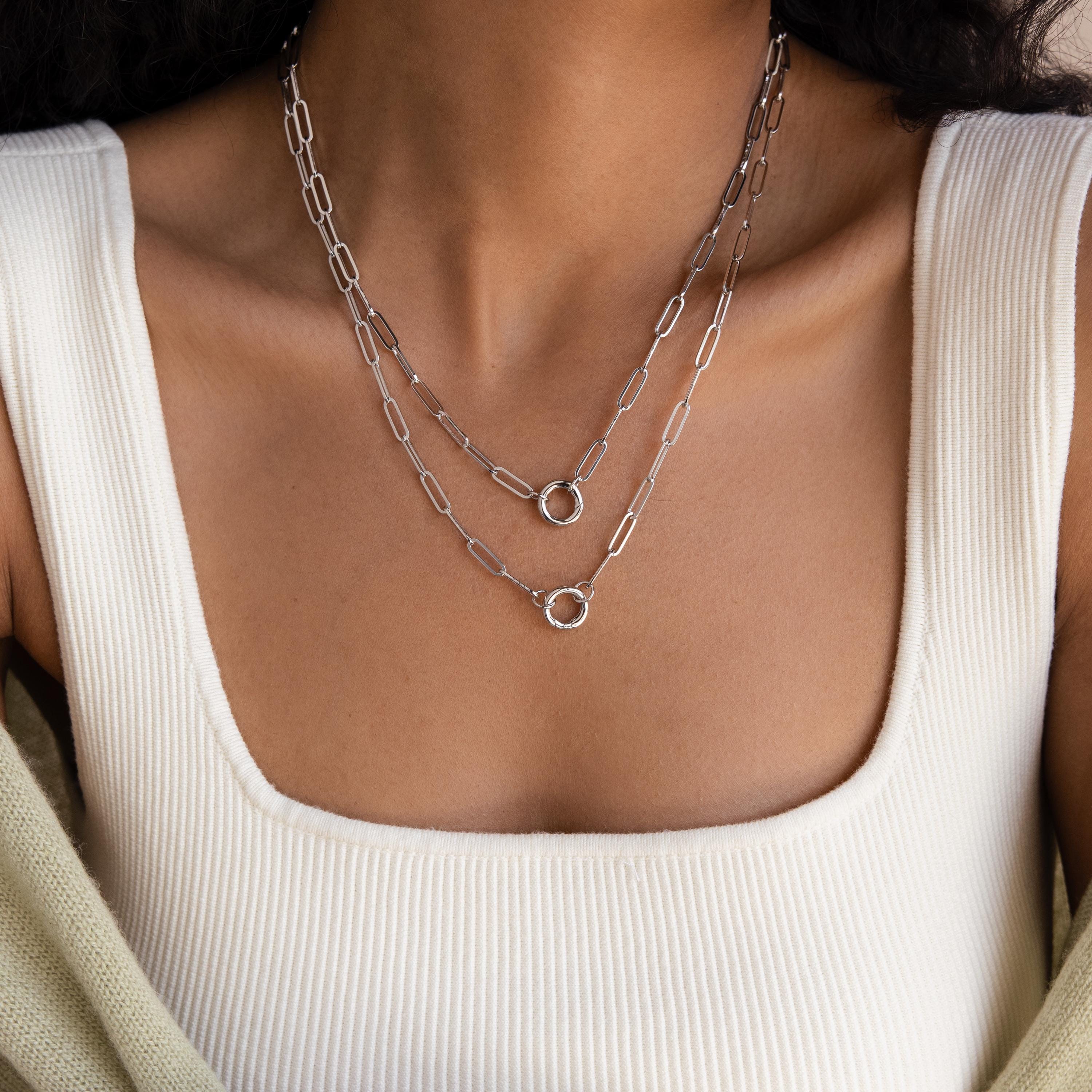 Close-up of a woman in a white square-neck top wearing the Paperclip Clasp Necklace—a layered silver chain featuring bold links, collectible charms, and circular pendants.
