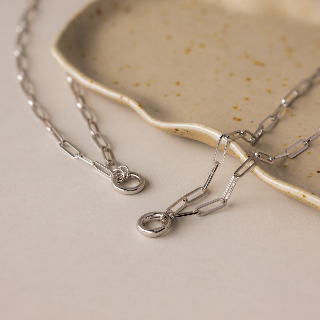 The Paperclip Clasp Necklace, a bold silver chain, rests on a beige speckled ceramic dish—ideal for pairing with your favorite collectible charms.