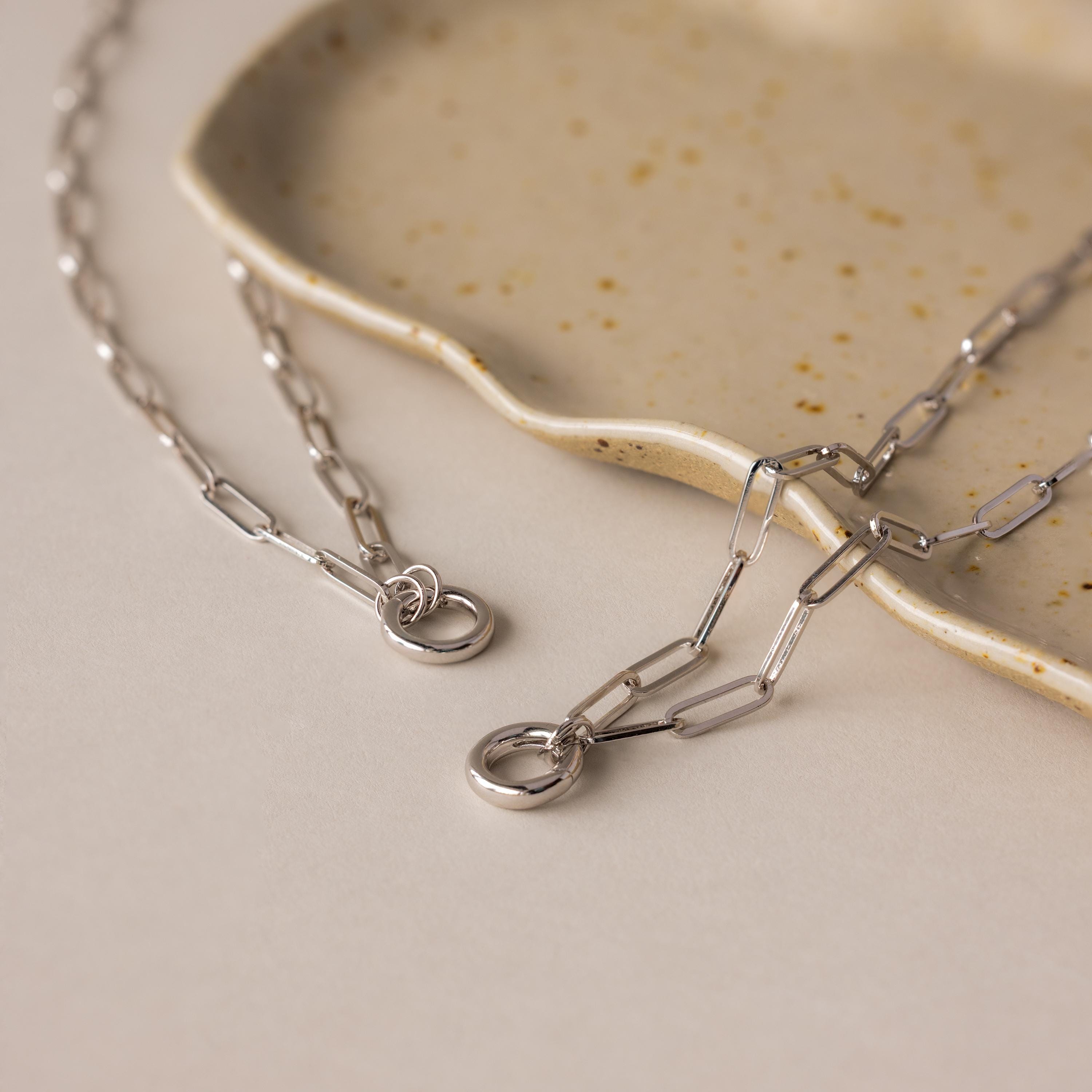 The Paperclip Clasp Necklace, a bold silver chain, rests on a beige speckled ceramic dish—ideal for pairing with your favorite collectible charms.