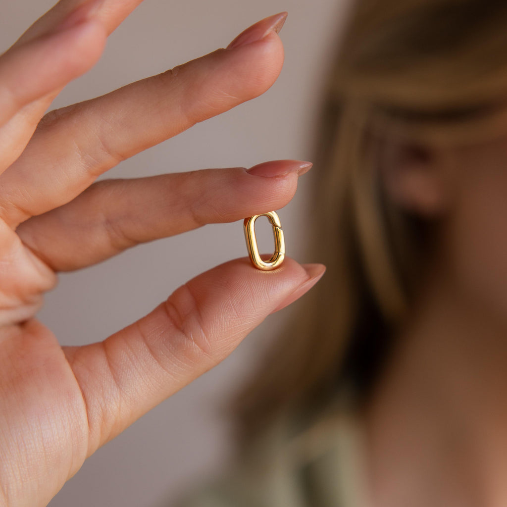 A hand with manicured nails holds the Multipurpose Charm Hoop Clasp Set in 18K Gold, oval-shaped and elegant, near a softly blurred face.