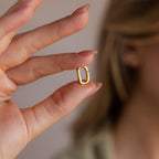 A hand with manicured nails holds the Multipurpose Charm Hoop Clasp Set in 18K Gold, oval-shaped and elegant, near a softly blurred face.