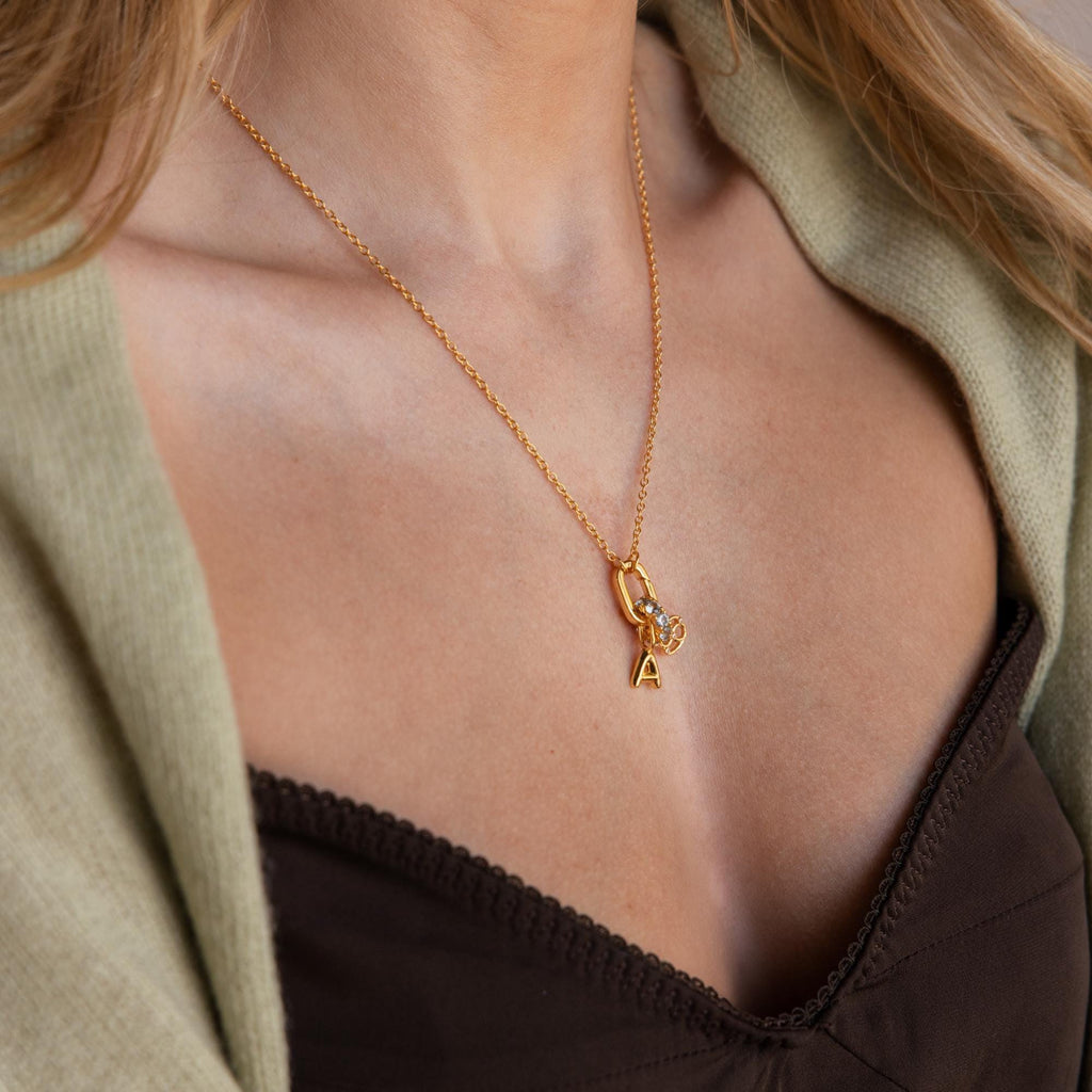 A woman wears the Multipurpose Charm Hoop Clasp Set in 18K Gold, styled with a letter A pendant and crystal charm, layered over a green cardigan and black top, creating a chic look with stacked bracelets.