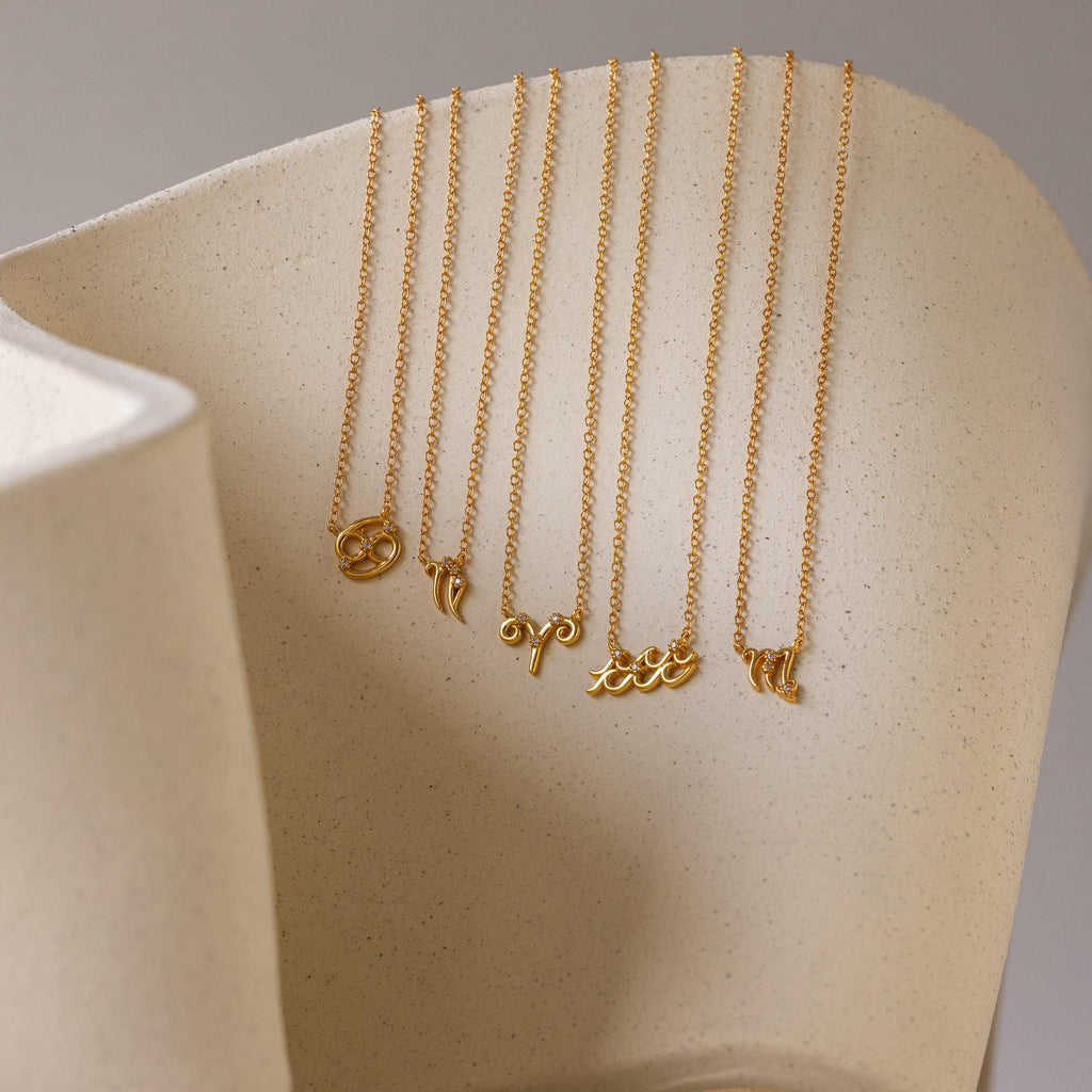 Five gold dainty zodiac charm necklaces hanging from a ceramic dish. Zodiacs in order: Cancer, Capricorn, Aries, Aquarius, and Virgo.