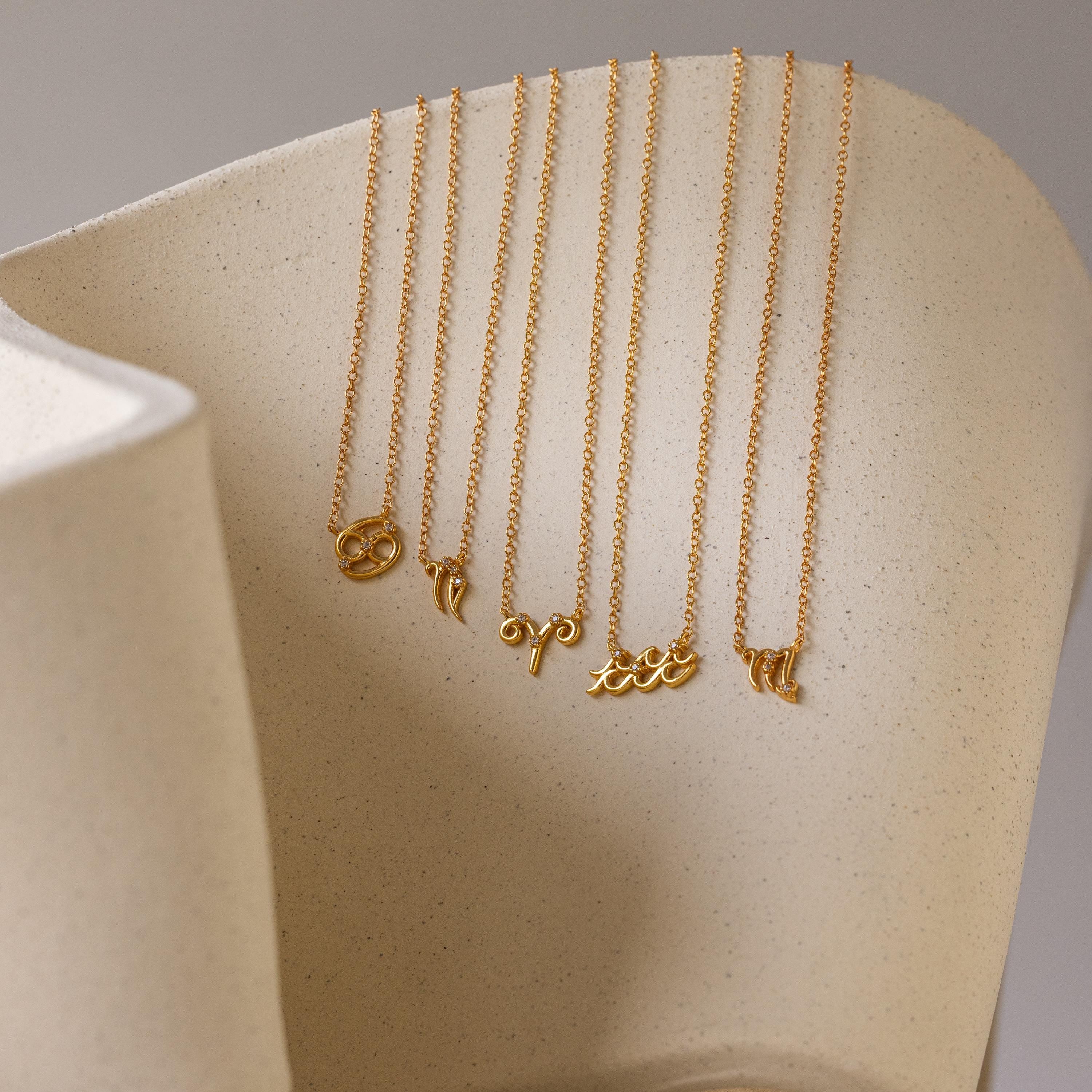 Five gold dainty zodiac charm necklaces hanging from a ceramic dish. Zodiacs in order: Cancer, Capricorn, Aries, Aquarius, and Virgo.