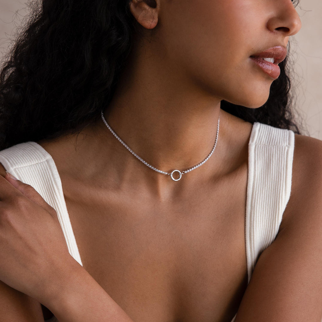 A woman wearing a white sleeveless top and the Diamond Tennis Clasp Necklace, featuring a small circular pendant and subtle rolo clasp, poses with her arms crossed over her chest.