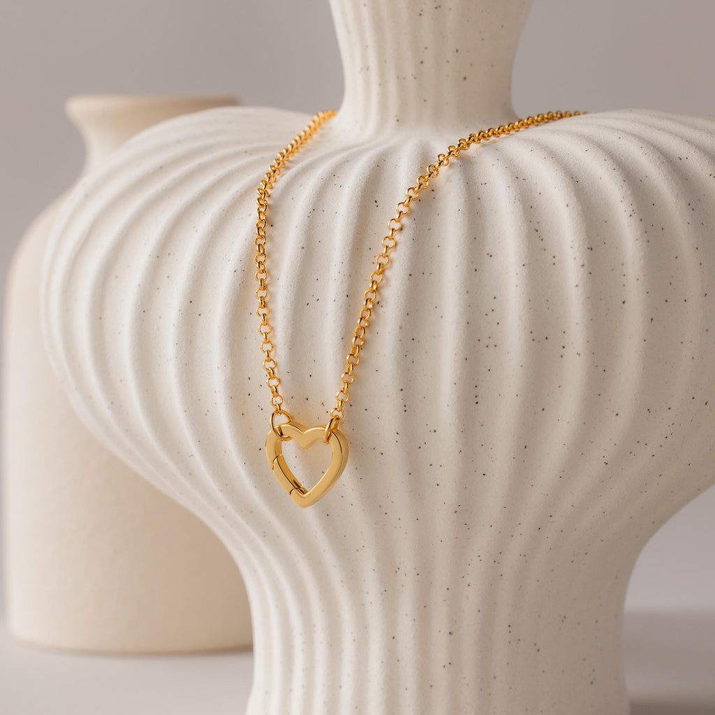 The Rolo Heart Clasp Necklace with a heart-shaped clasp is draped over a textured white ceramic vase, with another vase visible in the background.