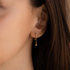 Model wears Gold Star Drop Chain Earrings. The design features a gold hoop earring with a star shaped charm and diamond in the center. A short dangling chain hangs from the hoop with another star shaped charm and diamond at the end.