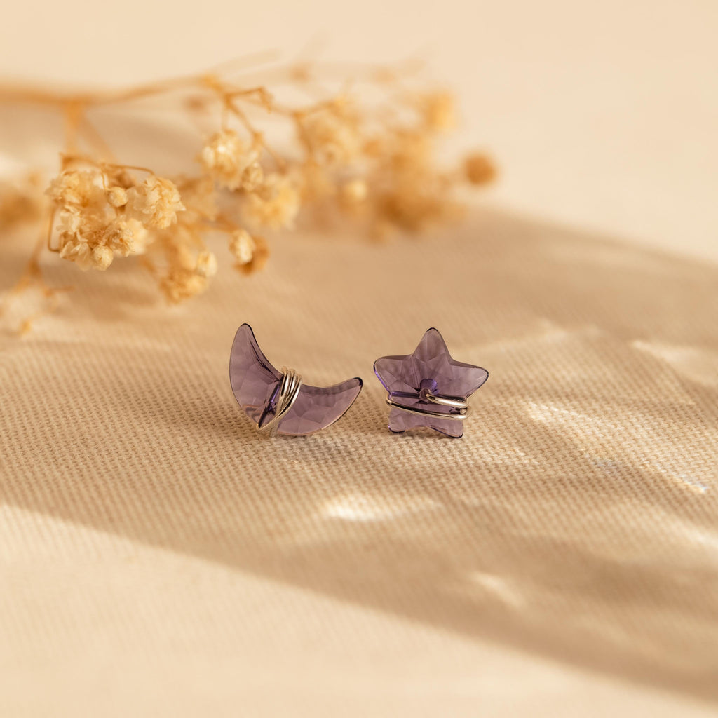Purple moon and star earrings in silver sit on a beige fabric with dried flowers in the background.