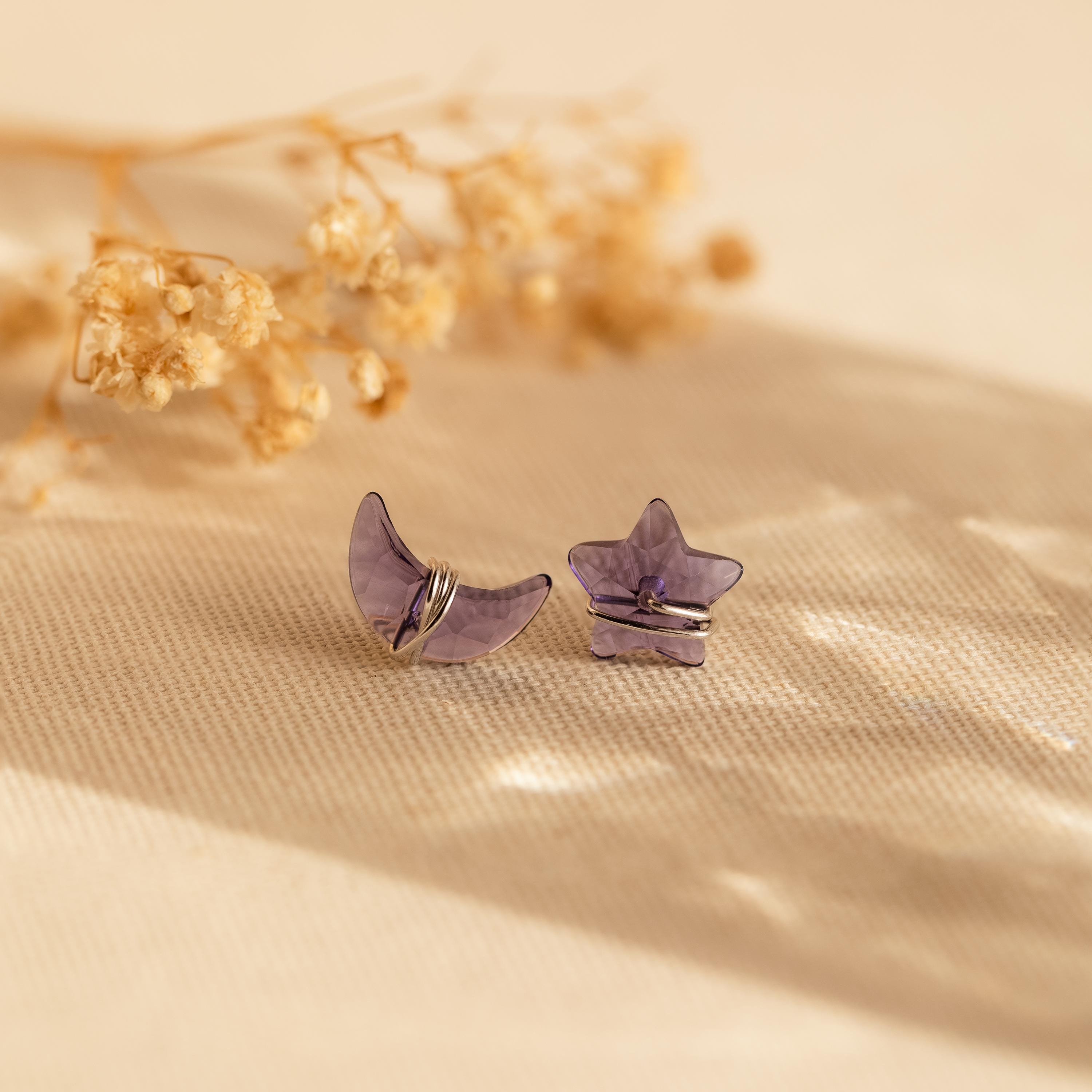 Purple moon and star earrings in silver sit on a beige fabric with dried flowers in the background.