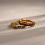 The Staggered Marquise Onyx Ring, featuring leaf-shaped green and red gemstones, elegantly rests on a light, textured surface alongside a second gold ring.