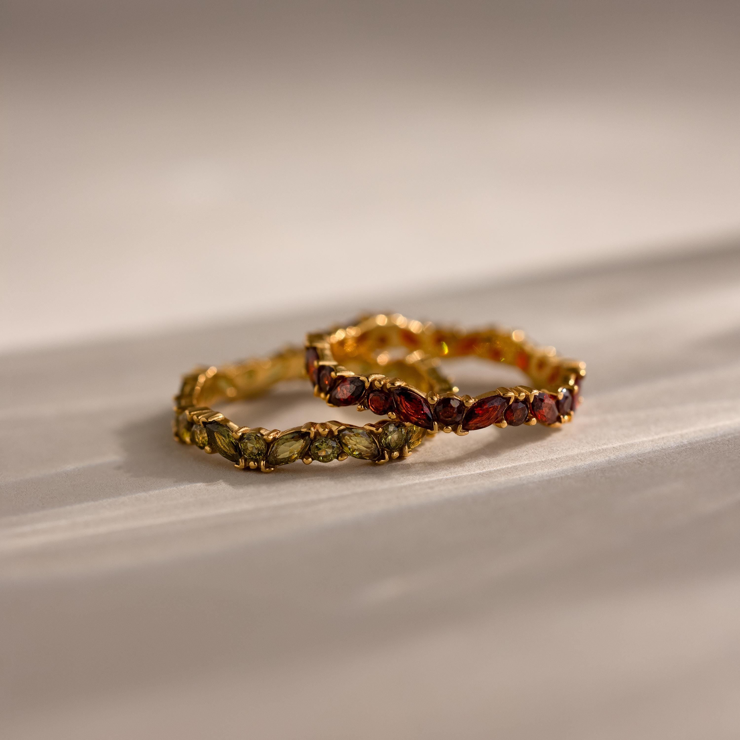 The Staggered Marquise Onyx Ring, featuring leaf-shaped green and red gemstones, elegantly rests on a light, textured surface alongside a second gold ring.