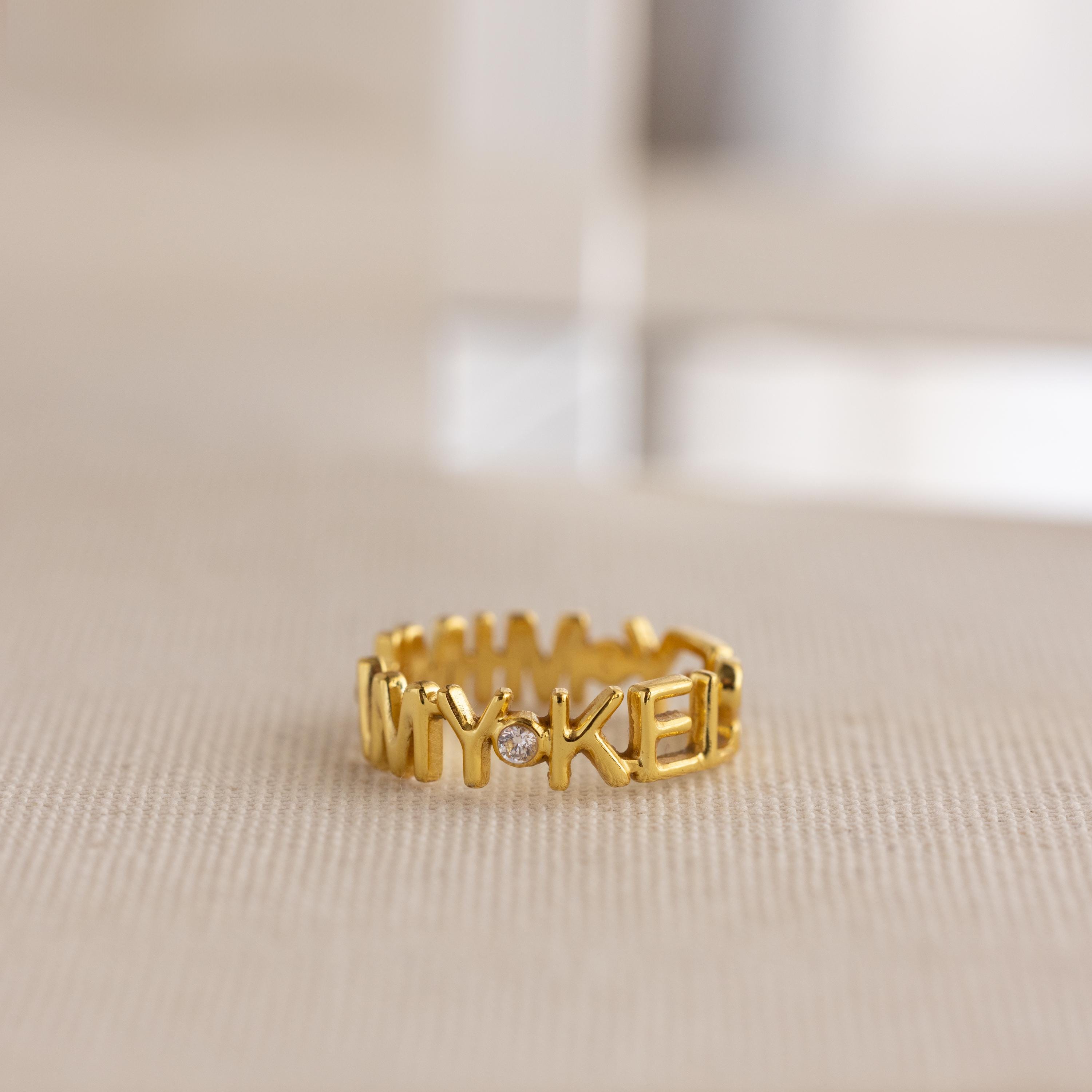 The Multiple Name Eternity Ring in 18K Gold features custom "MY-KEEP" engraving and a sparkling gemstone, displayed on beige fabric—inspired by timeless eternity ring designs with delicate diamond accents.
