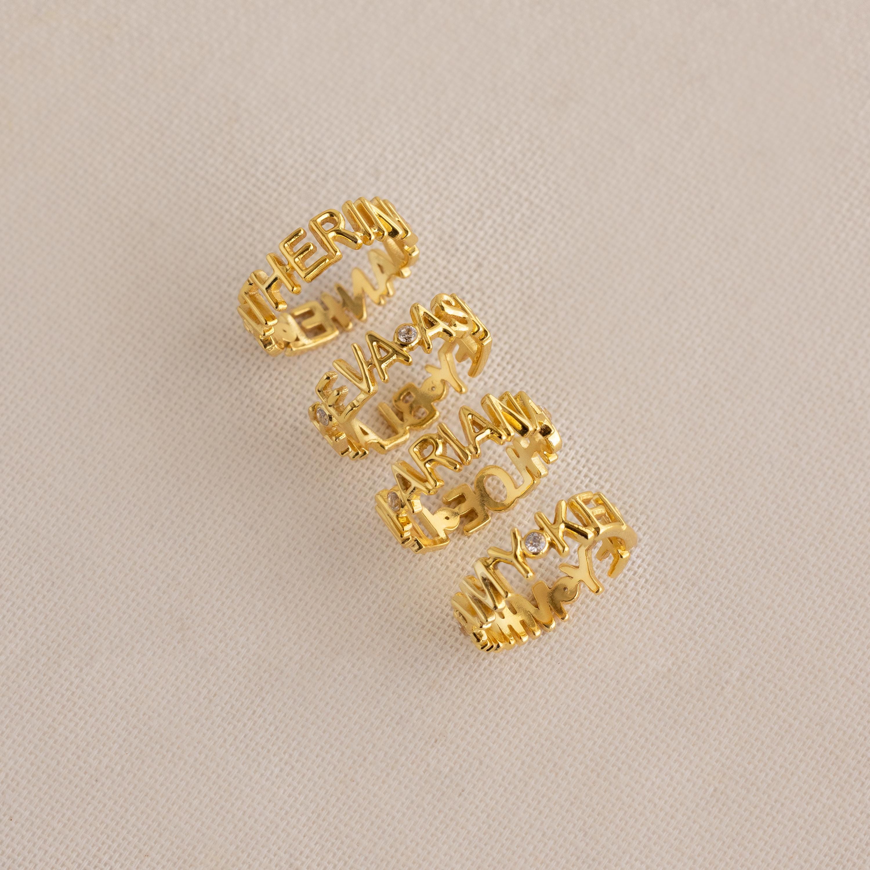 Four Multiple Name Eternity Rings in 18K Gold, each personalized with cut-out names, are arranged vertically on a beige textured surface—a unique gift to celebrate your loved ones.