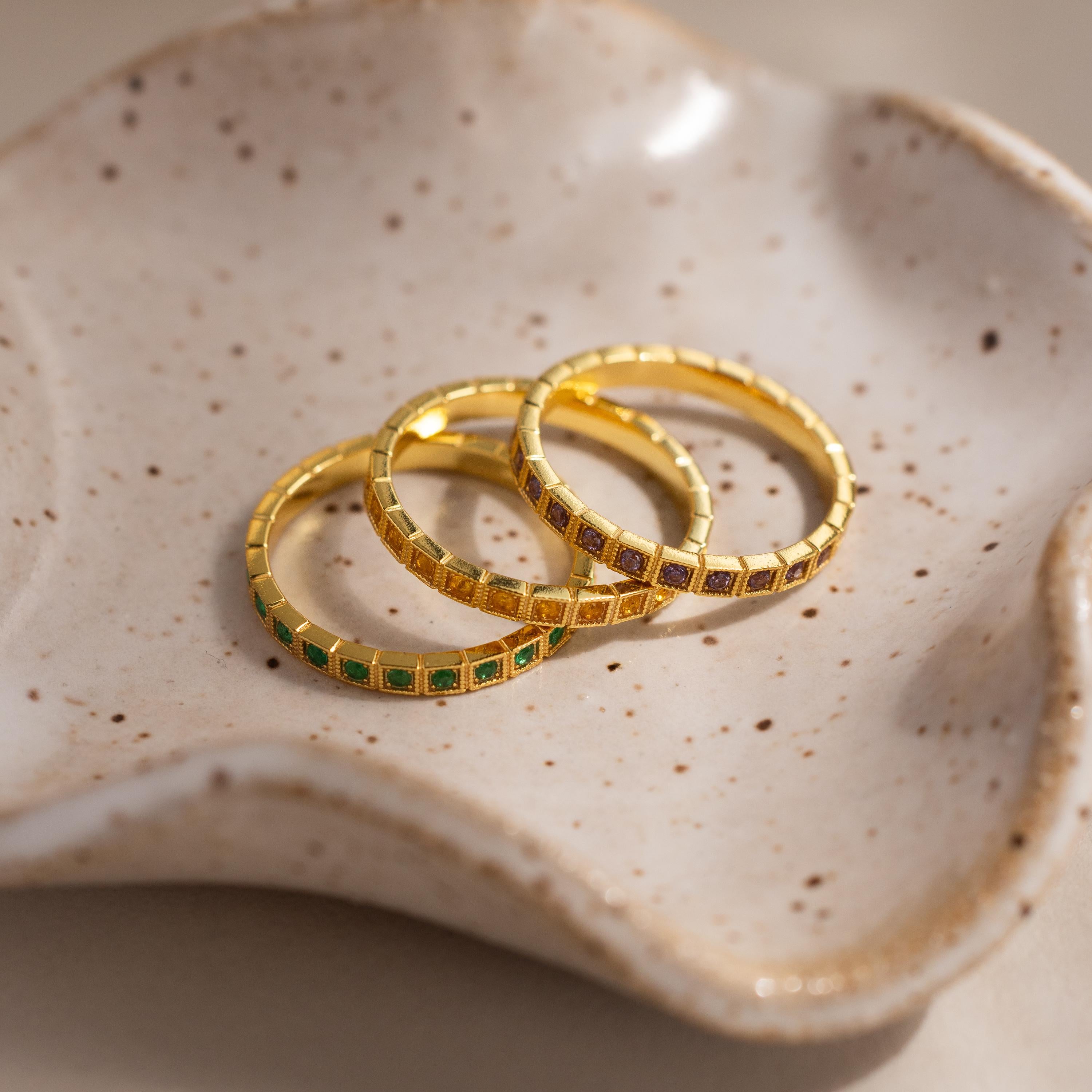 The Art Deco Emerald Eternity Ring, along with two other gold rings, rests on a speckled, wavy ceramic dish.