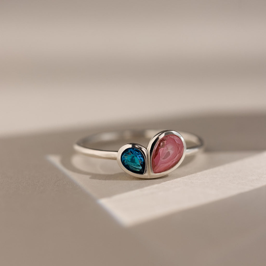 The Toi et Moi Heart Birthstone Ring in Sterling Silver, featuring blue and pink heart-shaped stones, rests on a light surface.