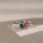 The Toi et Moi Heart Birthstone Ring in Sterling Silver, featuring blue and pink heart-shaped stones, rests on a light surface.
