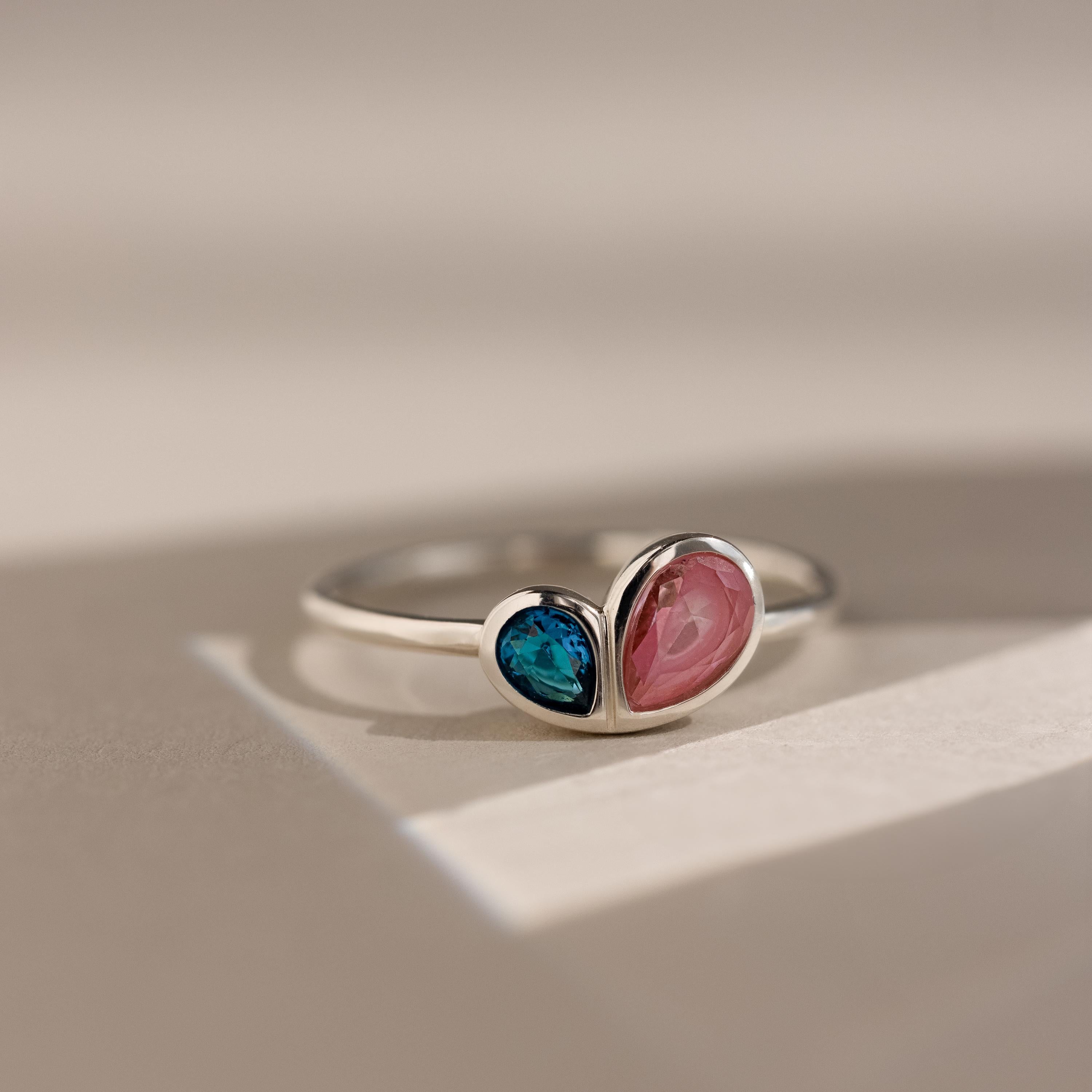 The Toi et Moi Heart Birthstone Ring in Sterling Silver, featuring blue and pink heart-shaped stones, rests on a light surface.