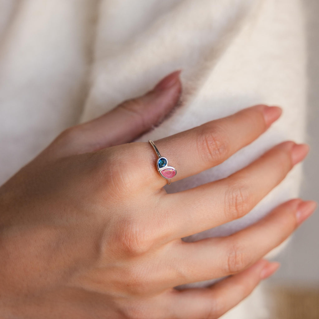 A manicured hand wears the Toi et Moi Heart Birthstone Ring in Sterling Silver, featuring blue and pink gemstones, against a soft white fabric backdrop.