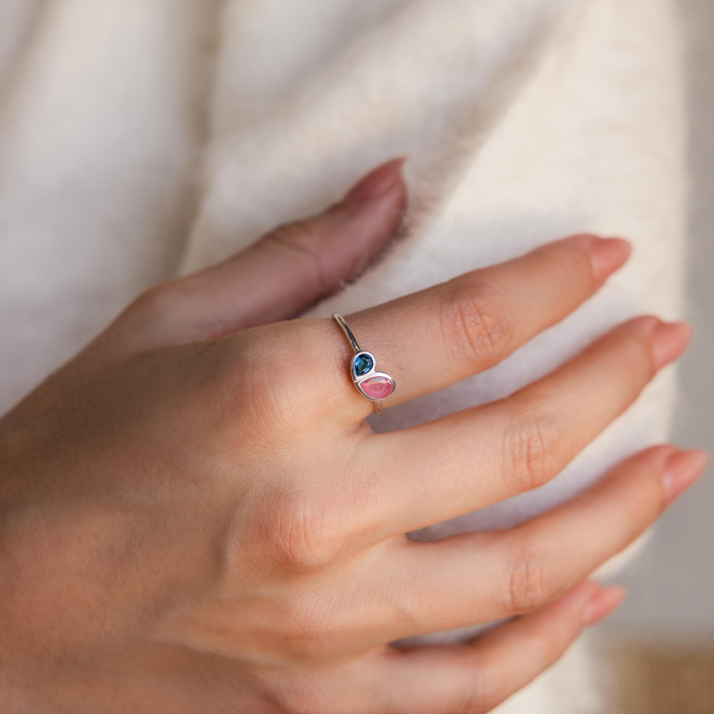 A manicured hand wears the Toi et Moi Heart Birthstone Ring in Sterling Silver, featuring blue and pink gemstones, against a soft white fabric backdrop.