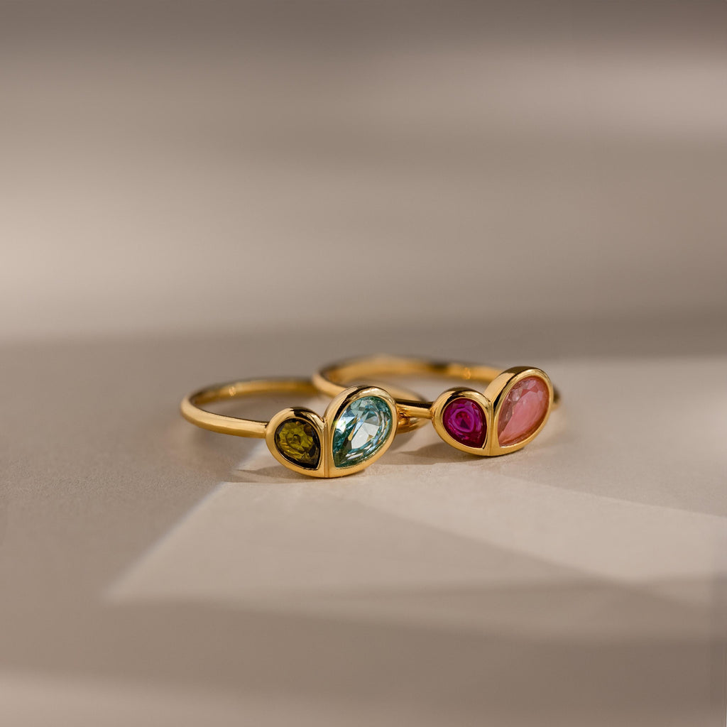 Two Toi et Moi Heart Birthstone Rings, each with heart-shaped settings and two differently colored pear-cut gemstones, are displayed on a soft background.