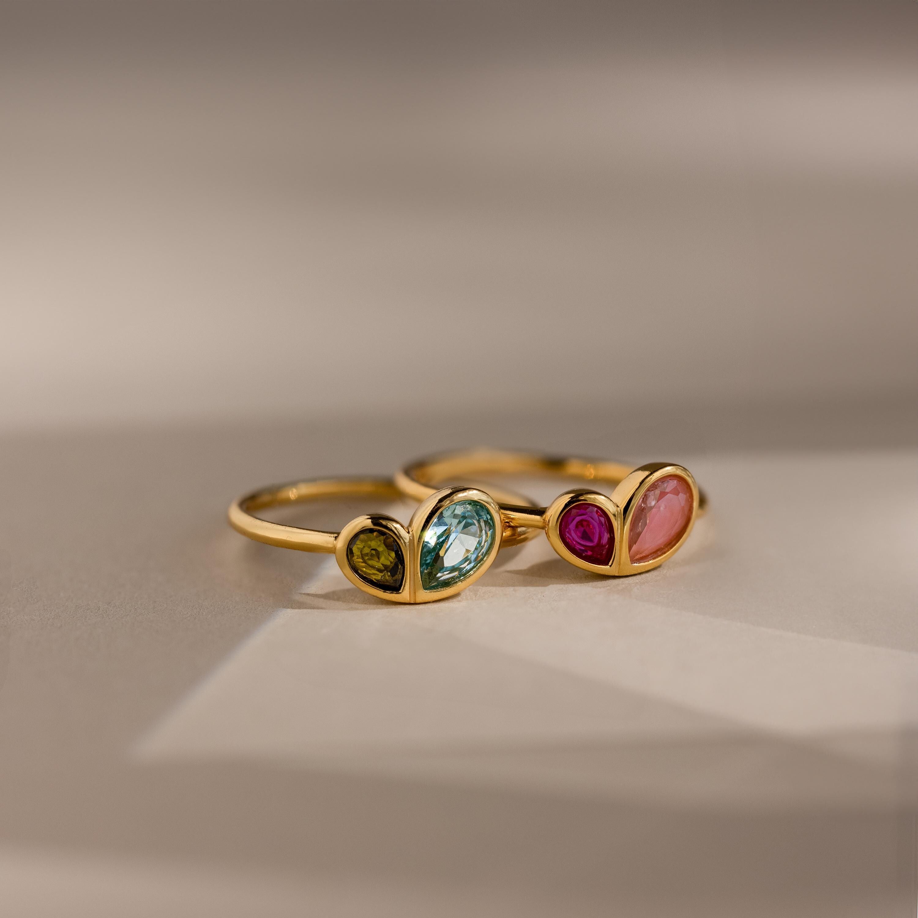 Two Toi et Moi Heart Birthstone Rings, each with heart-shaped settings and two differently colored pear-cut gemstones, are displayed on a soft background.