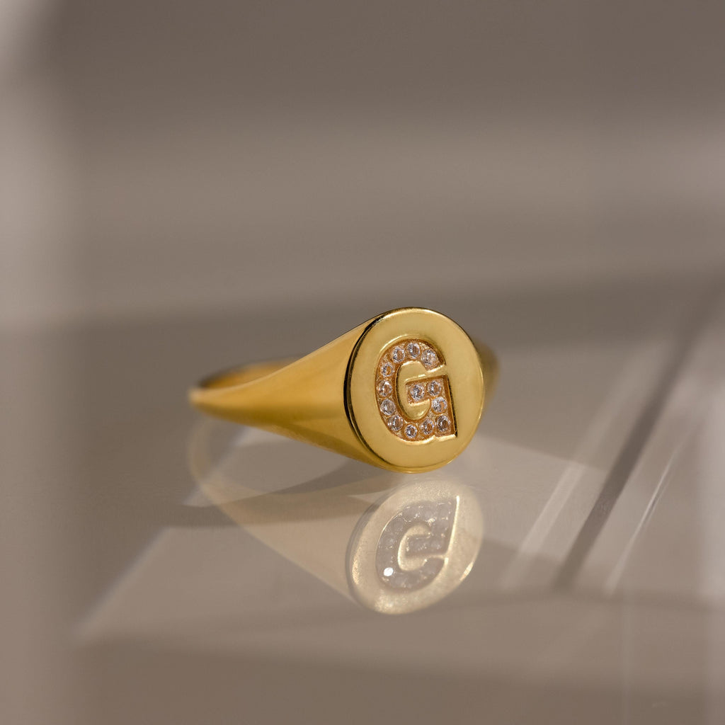 The Pave Initial Signet Ring in 18K Gold features a crystal-studded letter G engraved on its round face, reflecting light beautifully—an eye-catching statement piece.
